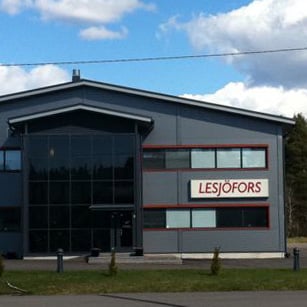 Mūsdienīga rūpnieciskā ēka ar Lesjöfors zīmi, lieliem logiem un elegantu dizainu. Tā atrodas uz koku fona zem daļēji mākoņainām debesīm.