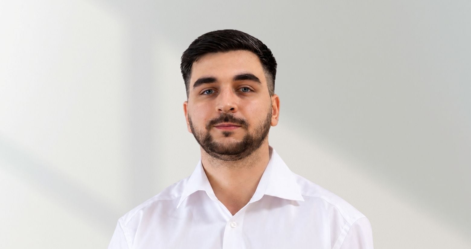 Emre Inci steht vor einer weißen Wand und trägt ein weißes Hemd. Er hat schwarze glatte Haare und einen schwarzen Bart.
