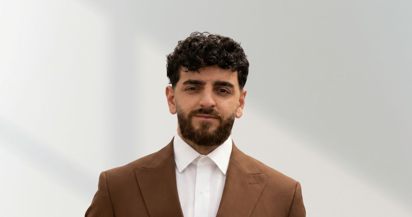 Hakan Güler steht vor einer weißen Wand und trägt einen braunen Anzug. Er hat schwarze lockige Haare und einen schwarzen Bart.
