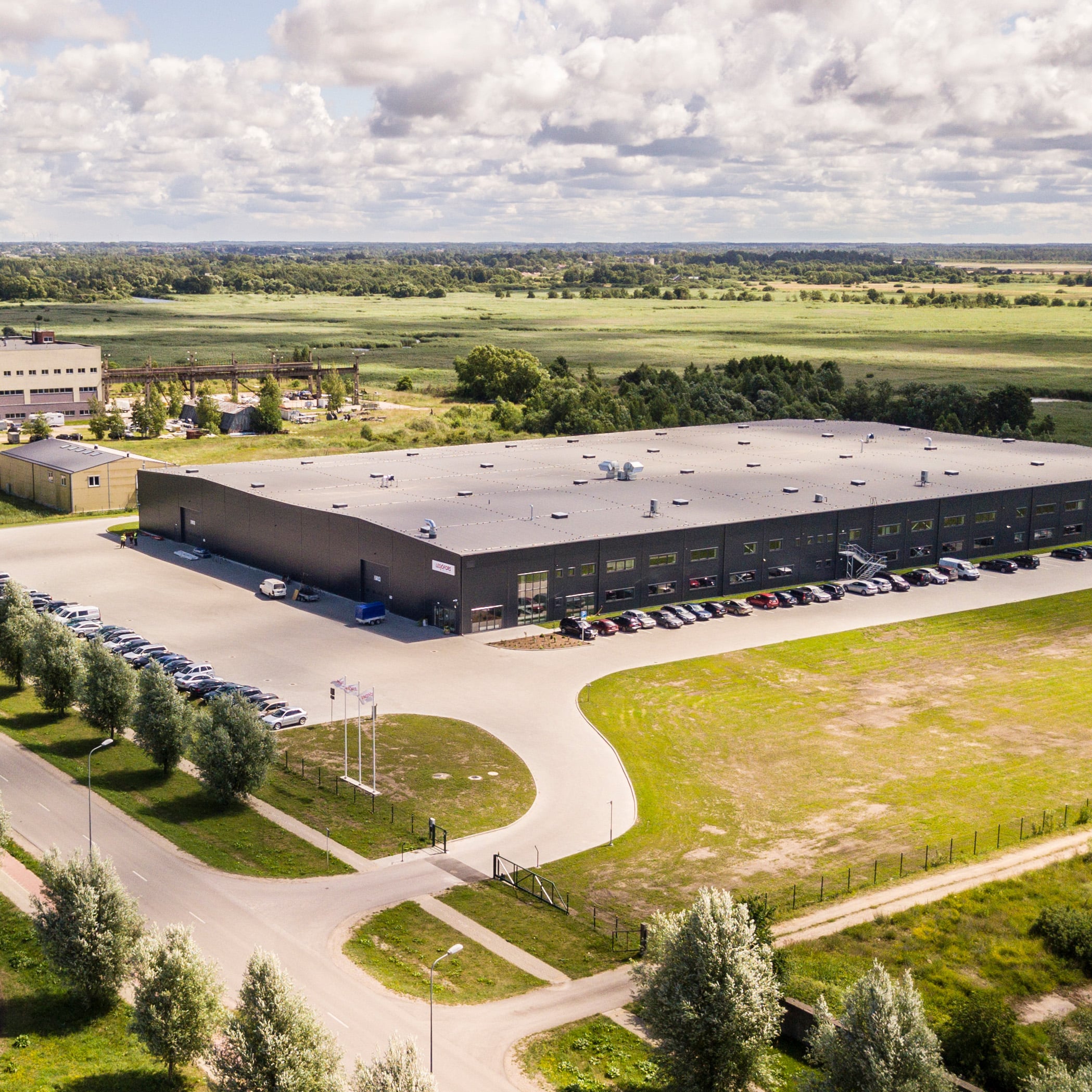 Aerofotogrāfija ar lielu rūpniecisko ēku, ko ieskauj autostāvvietas un koki. Plakana ainava stiepjas līdz horizontam zem daļēji mākoņainām debesīm.