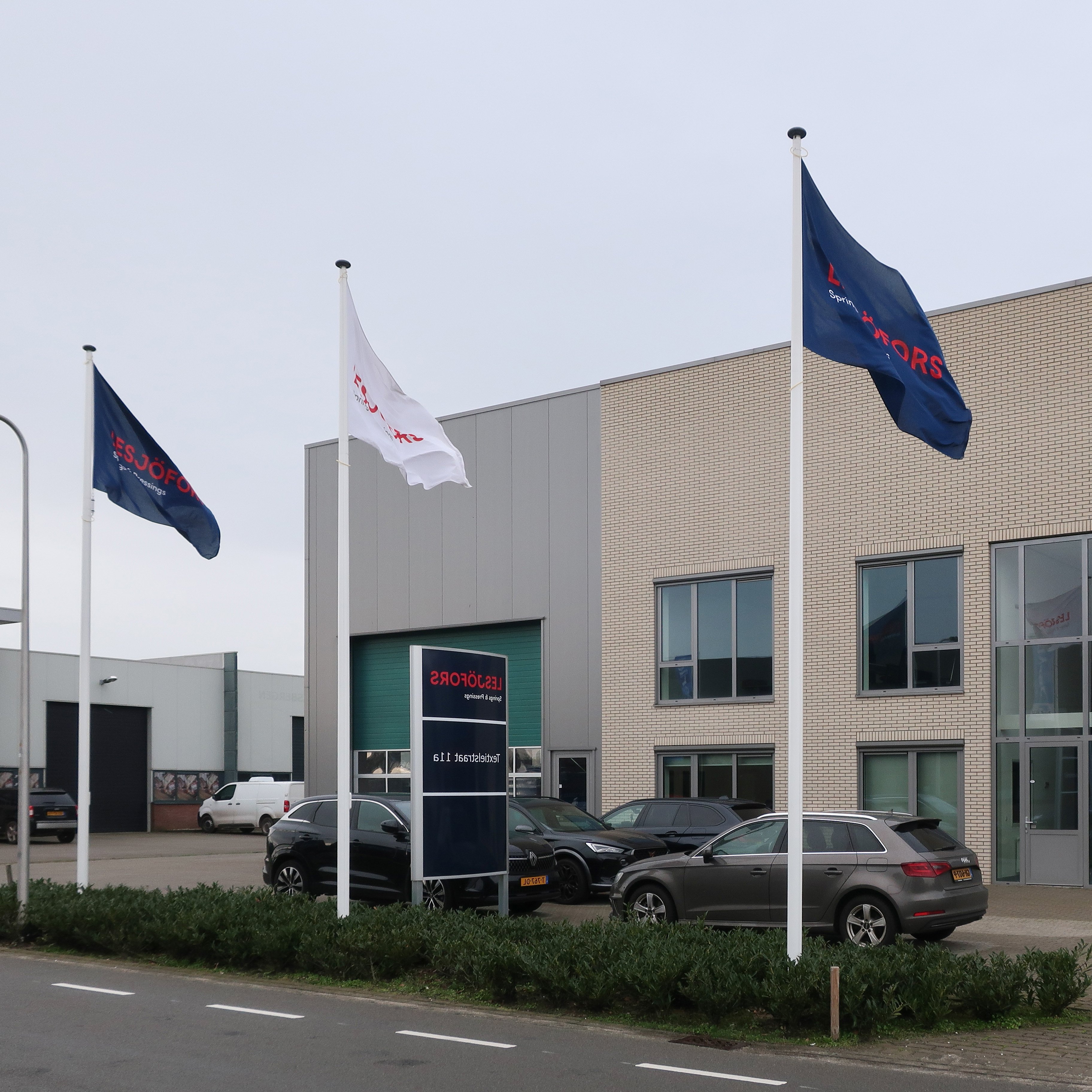 Contemporary industrial building with beige brick facade, large windows, and three flags in front. Overcast sky and parked cars on a paved area.