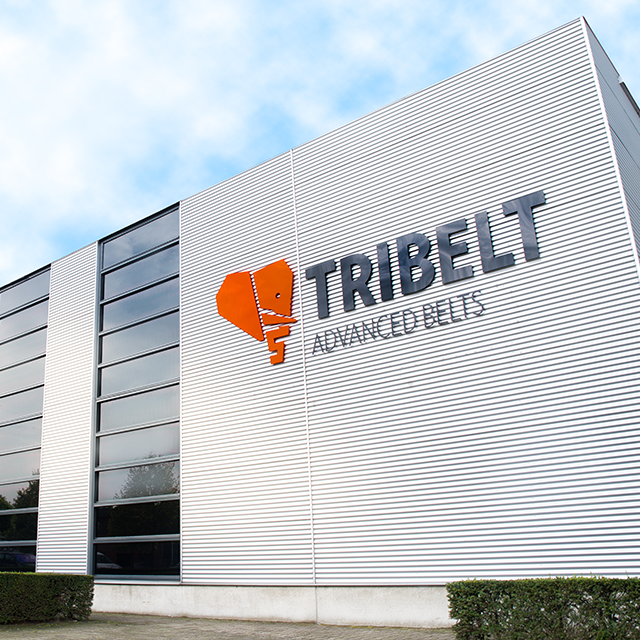 Mūsdienīga rūpnieciskā ēka ar atstarojošu stiklu un logotipu ar uzrakstu “Tribelt”. Koptie krūmi un skaidras zilas debesis piešķir profesionālu noskaņu.