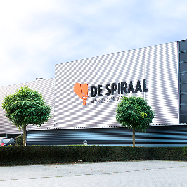 Mūsdienīga rūpnieciskā ēka ar logotipu “DE SPIRAAL” un oranžu sirdi. Divi koki un apgriezti dzīvžogi piešķir zaļumu. Debesis ir apmākušās.