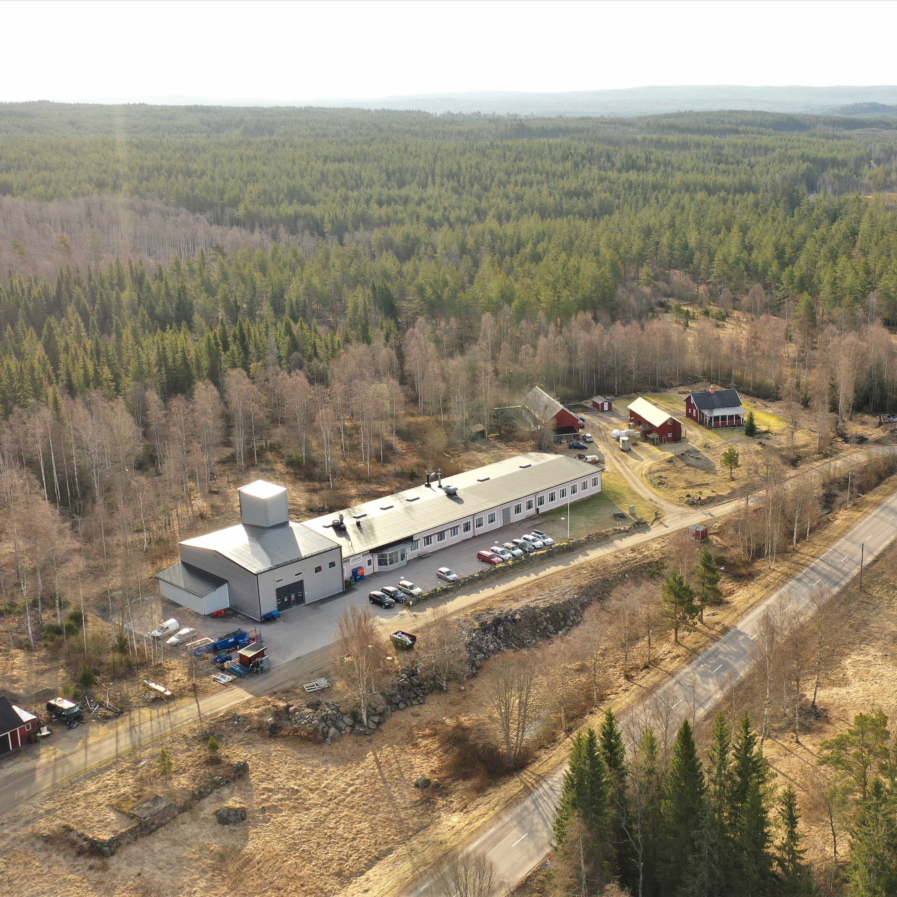 Aerial view of a rural landscape with a long, straight road cutting through a forest. A large industrial building and several red houses are nearby.
