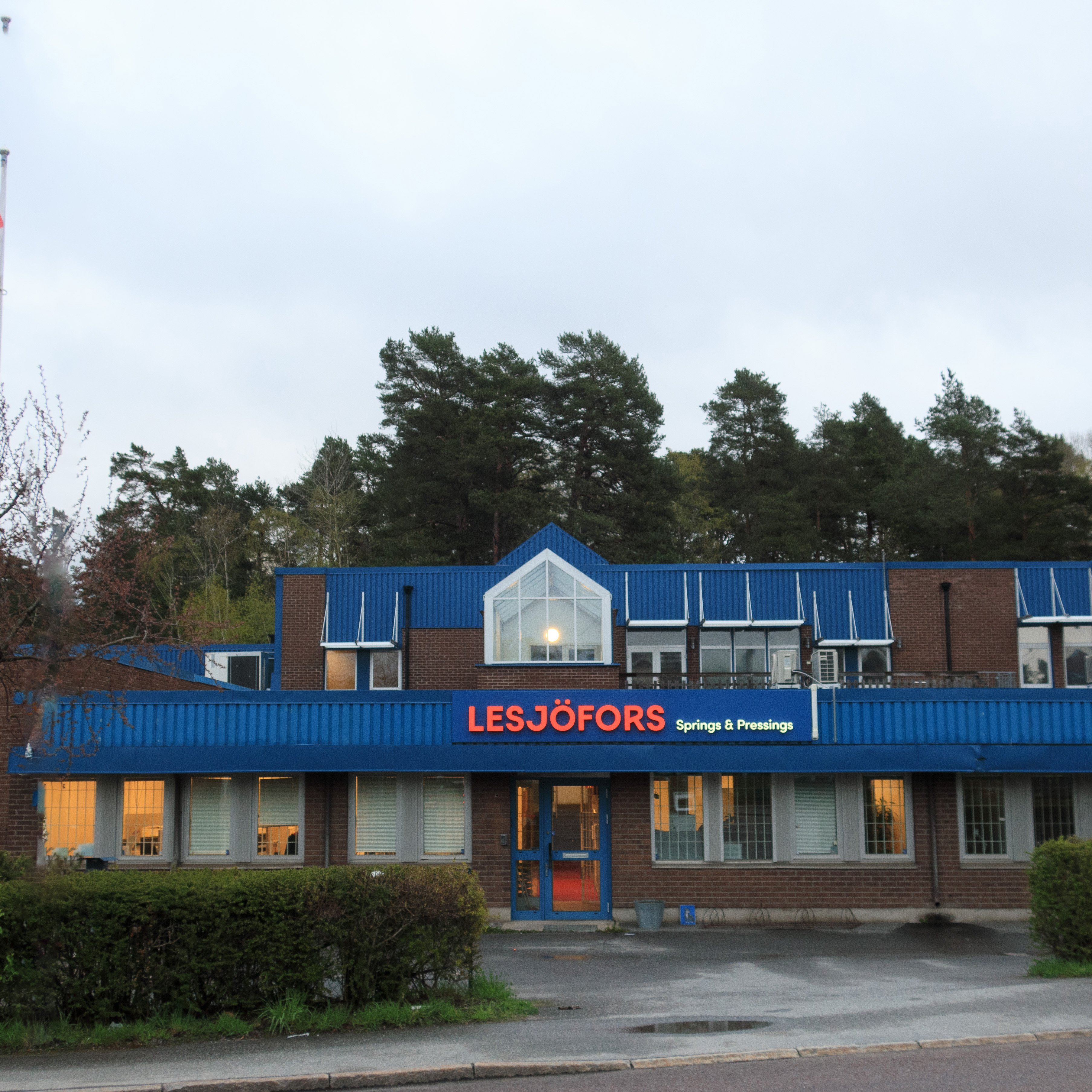 Ķieģeļu ēka ar zilu jumtu un uzrakstu “Lesjöfors Springs & Pressings”. To ieskauj koki, radot mierīgu un industriālu atmosfēru.