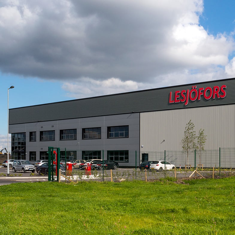 Stor industribygning med skiltet “Lesjöfors”, omgivet af en græsbevokset mark under en delvist skyet himmel.