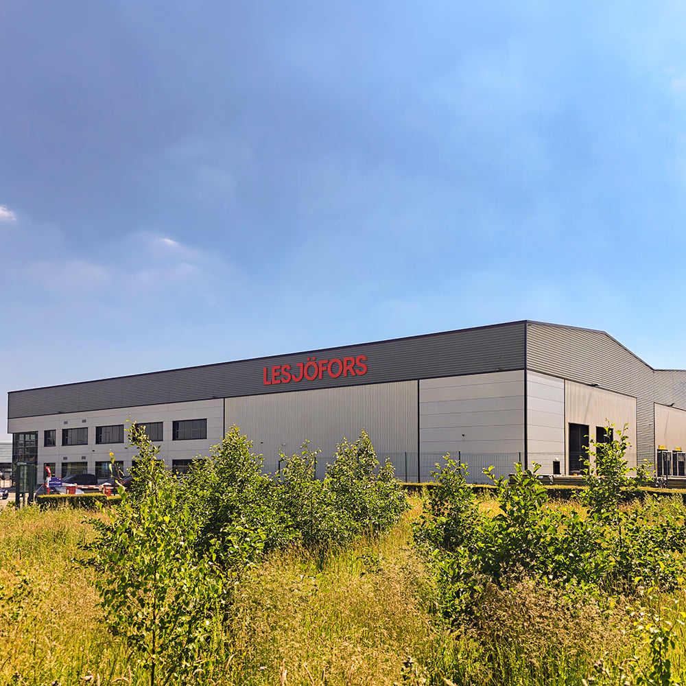 Eksteriør af Lesjöfors Springs UKs produktionsanlæg i Huddersfield, Storbritannien, med det røde Lesjöfors-logo på facaden og grøn beplantning foran bygningen.