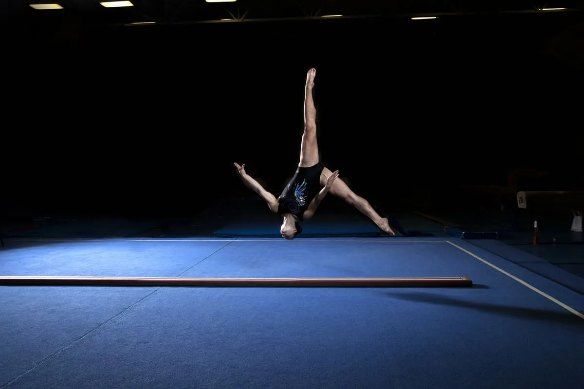 Gymnast performing an inverted handstand on a blue gymnastics floor inside a training gym.