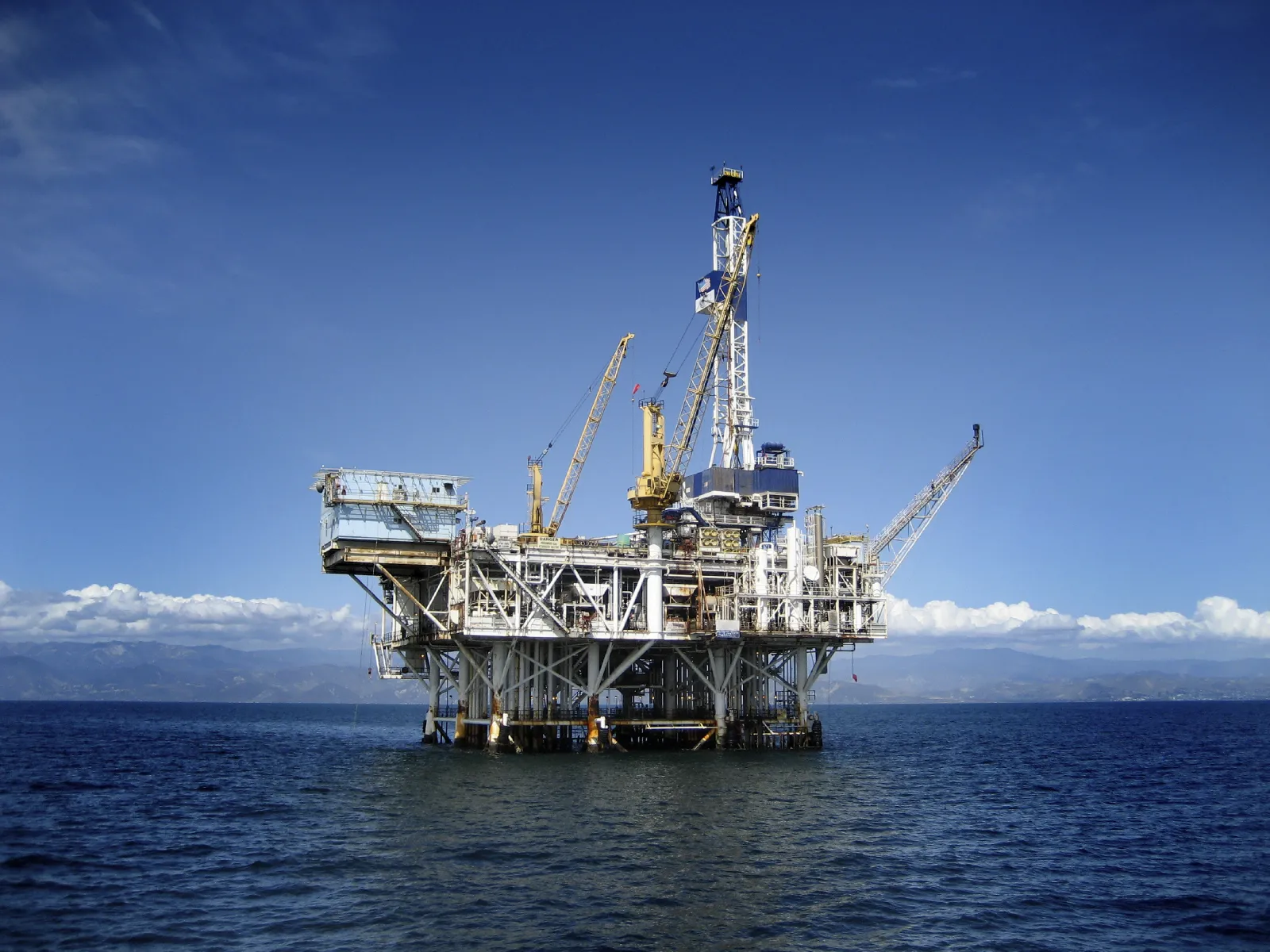 An offshore oil drilling platform standing on steel legs above calm blue ocean water, with mountains visible on the horizon under a clear blue sky