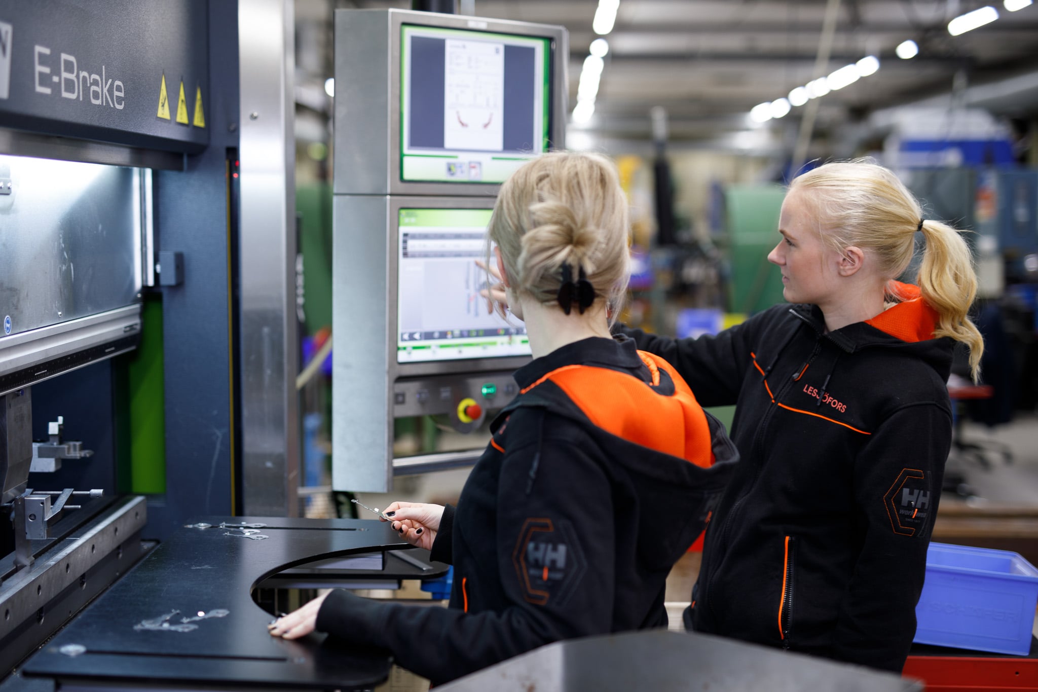Två arbetare i en fabrik, med svarta och orange uniformer, arbetar vid maskiner med digitala skärmar.