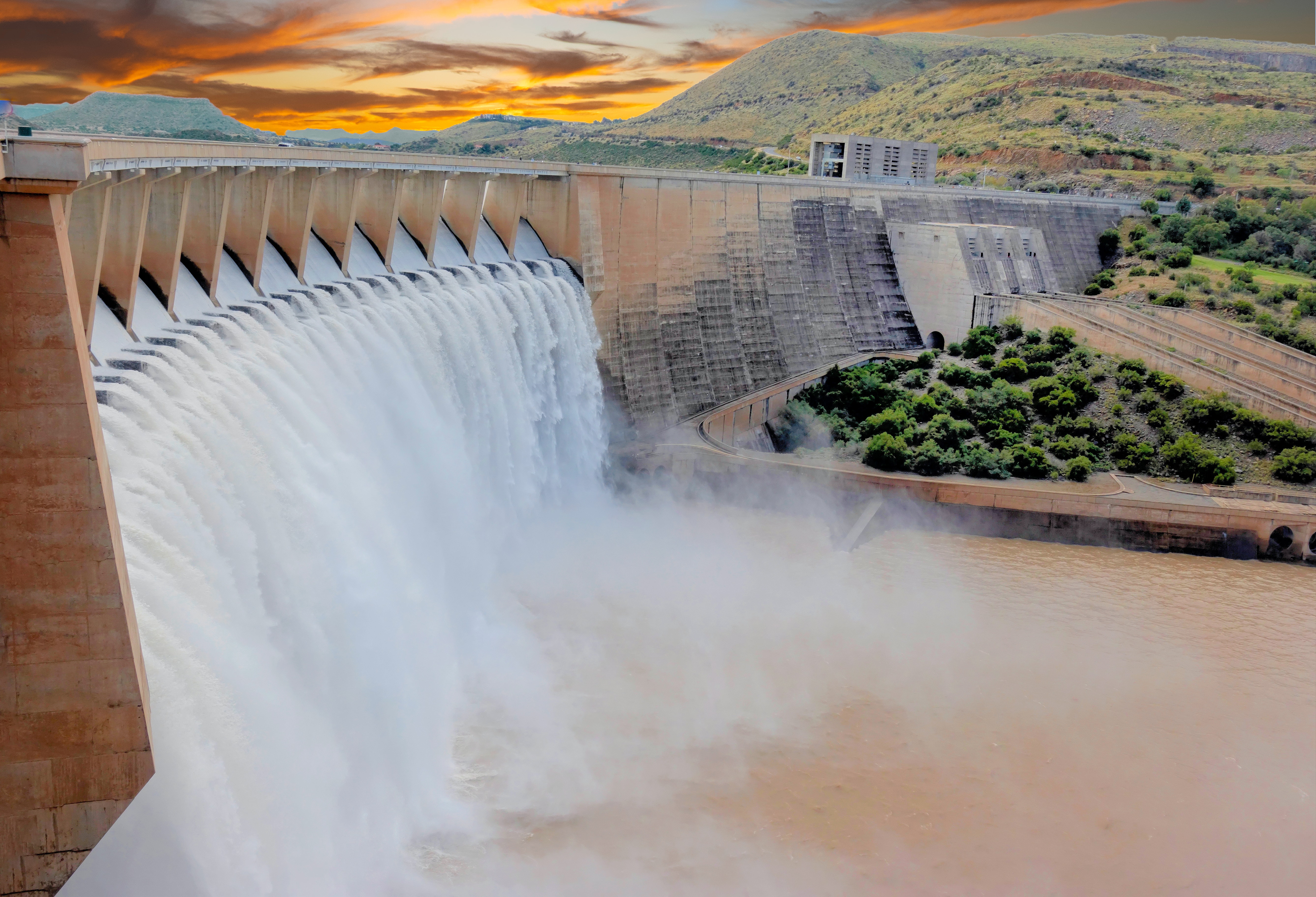 A large dam releases a powerful cascade of water into a river below, surrounded by lush greenery. The sky above is vibrant with a colorful sunset.