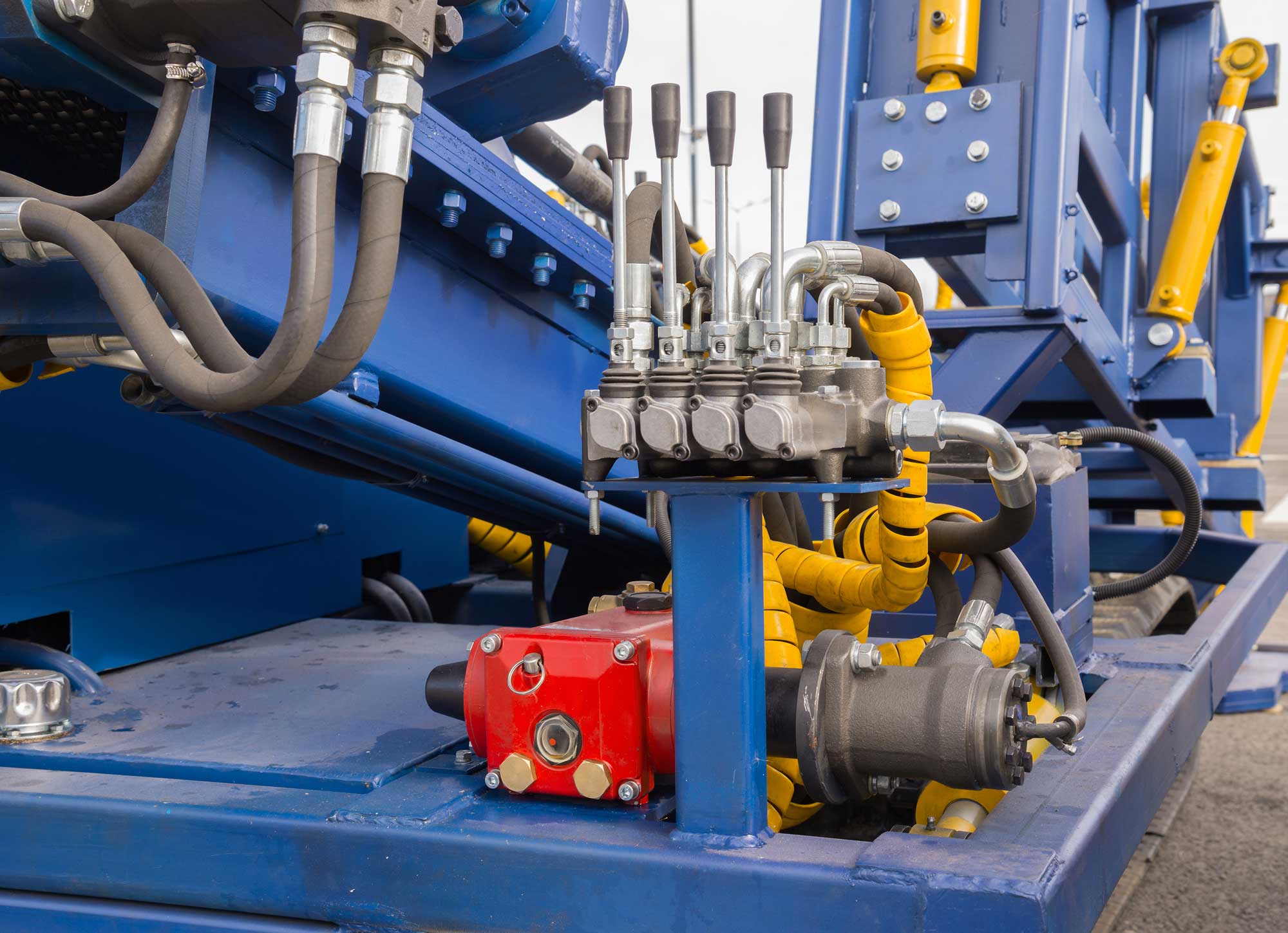 Nårbilde av industrielt hydraulmaskineri med blå og gule komponenter, som viser ventiler, rør og en rød kontrollboks.