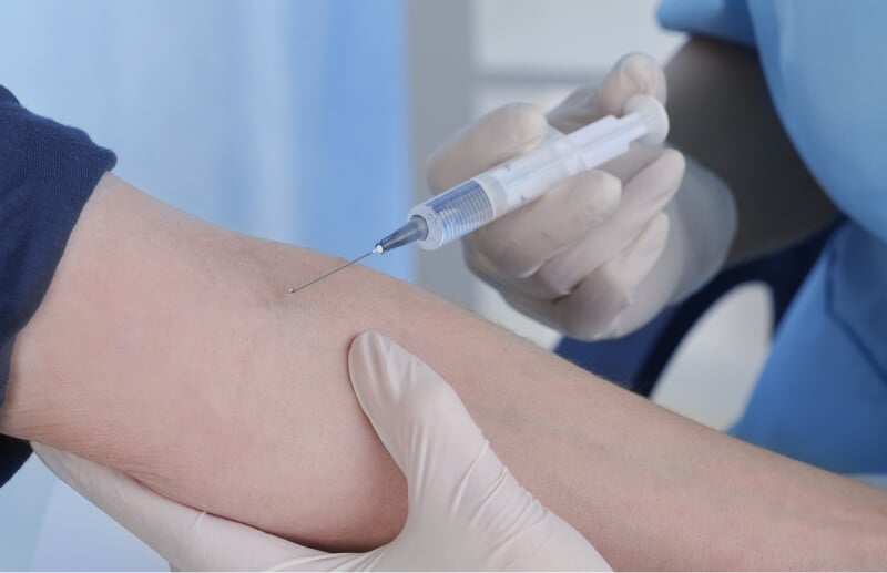 En sundhedsprofessionel i blåt arbejdstøj giver en injektion i en patients arm. Personen bærer handsker.