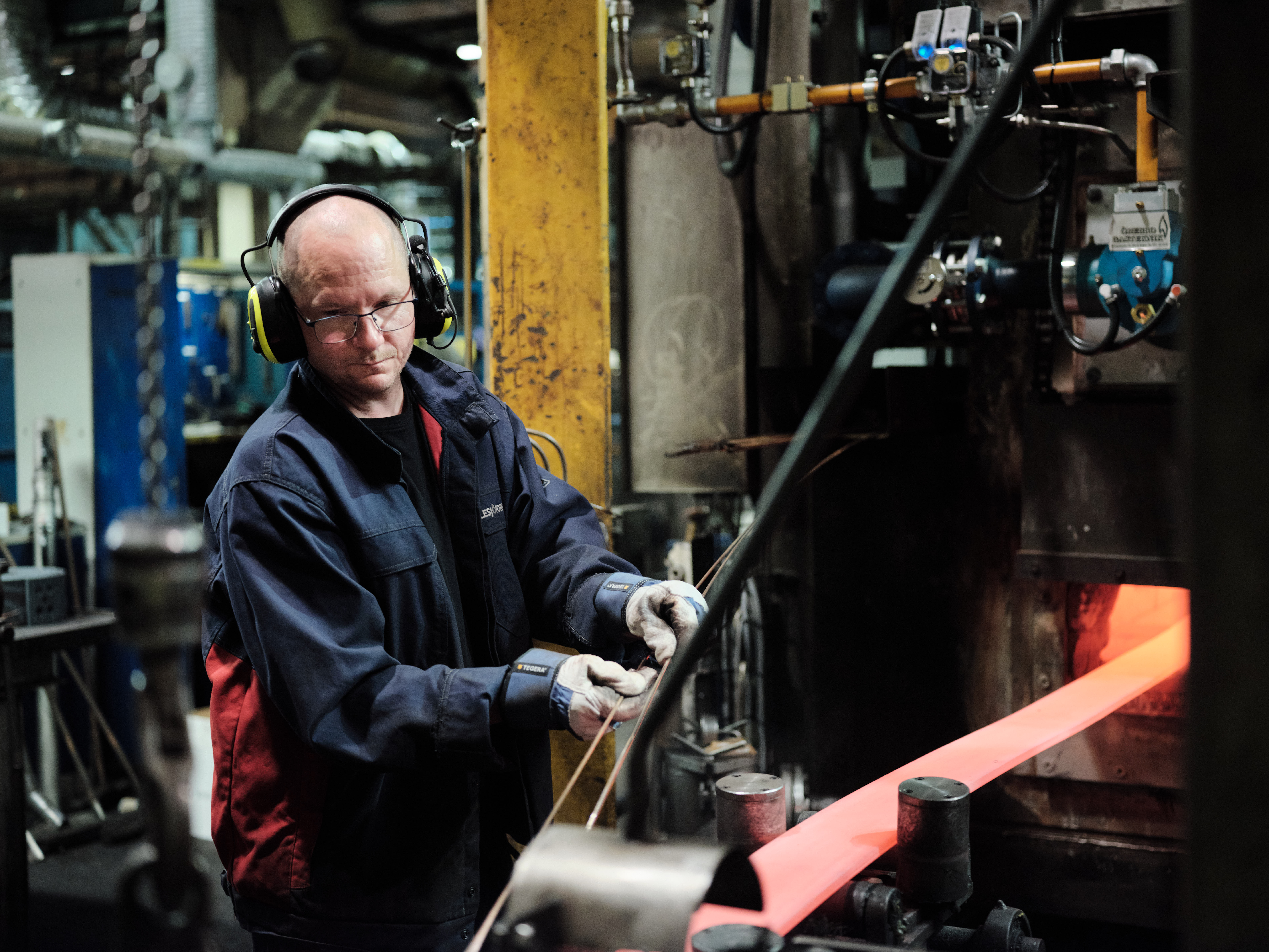 En arbeider i et verksted former en glødende metallstang ved hjelp av tungt maskineri. Han bruker hørselsvern og står blant industrielt verktøy, fokusert på oppgaven.
