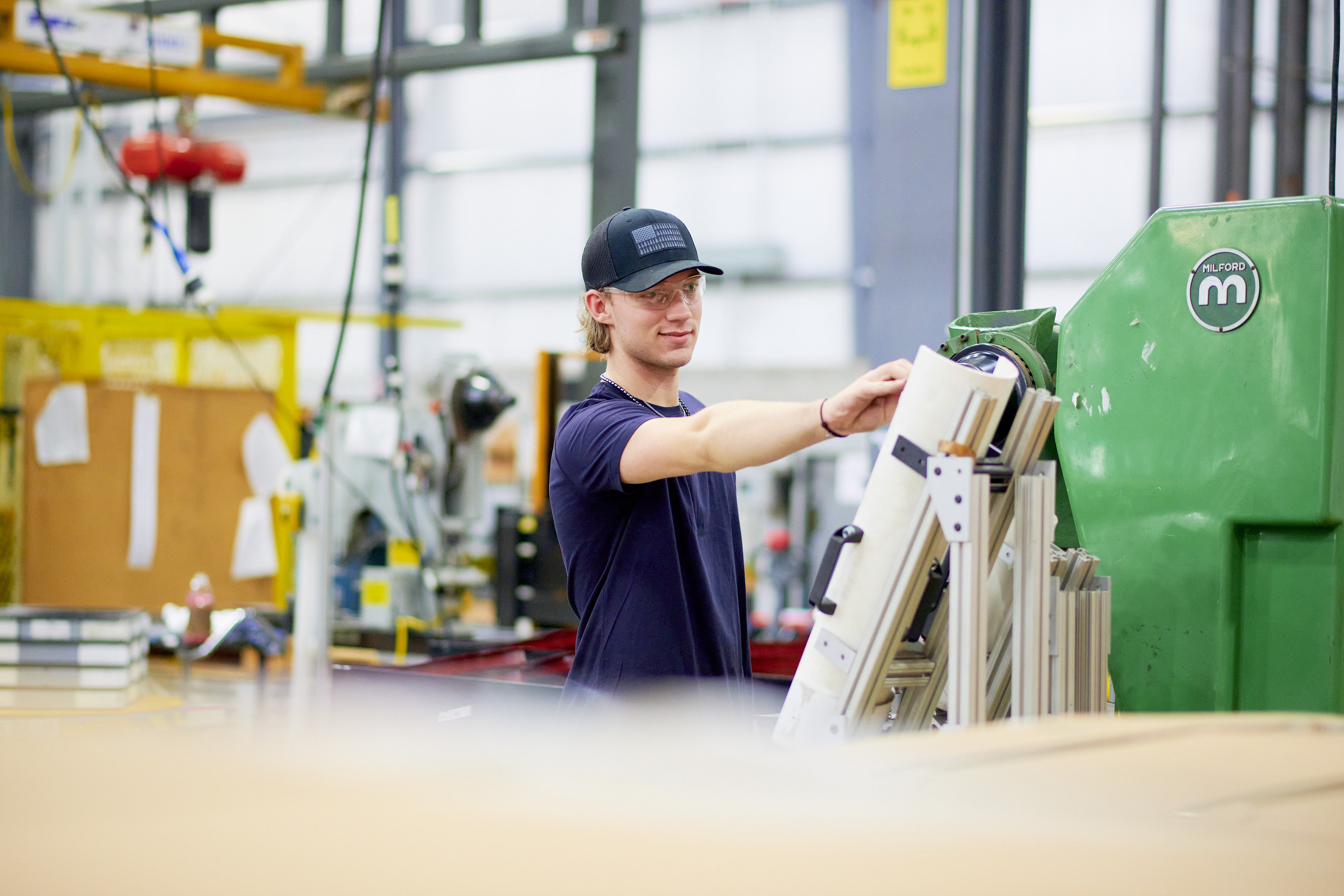En arbejder på en fabrik, iført kasket og marineblå skjorte, betjener maskineri. Miljøet er industrielt, med værktøj og udstyr synligt i baggrunden.