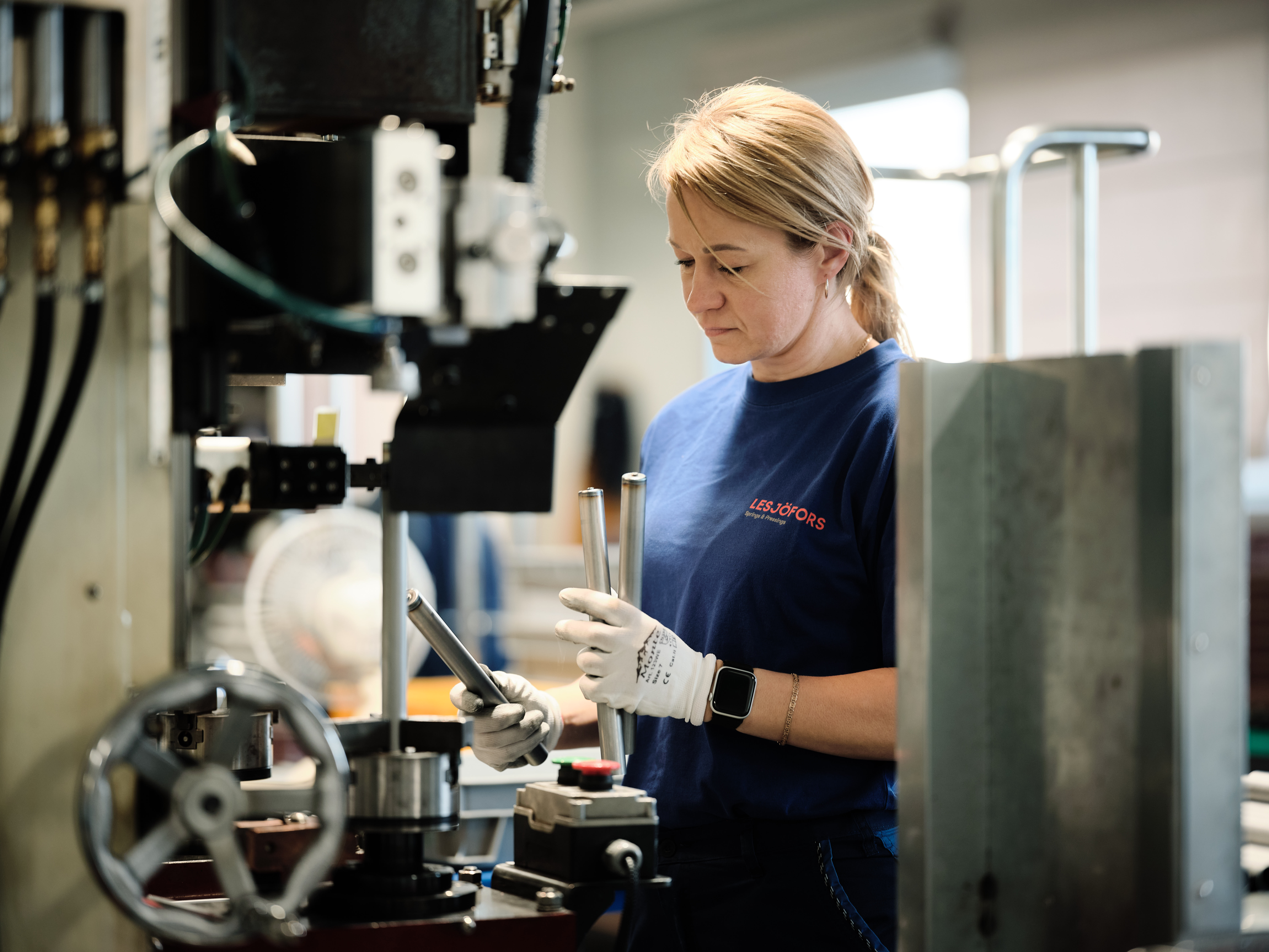 Eine Frau in einer Fabrikumgebung, mit blauem Shirt und Handschuhen, konzentriert sich auf die Bedienung von Maschinen.