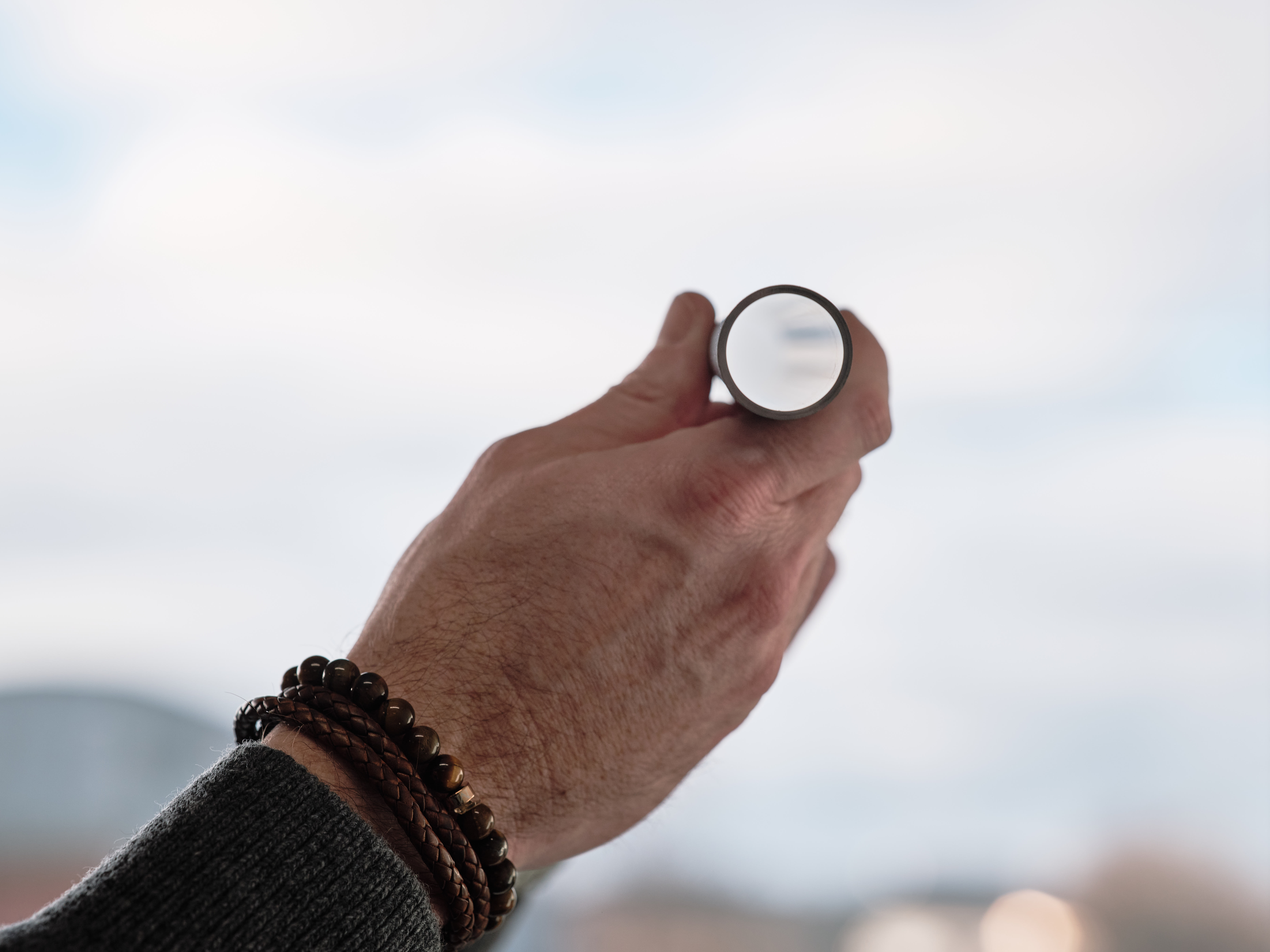 Eine Hand mit Armbändern hält eine Gasfeder in den Himmel, in der sich Wolken spiegeln.