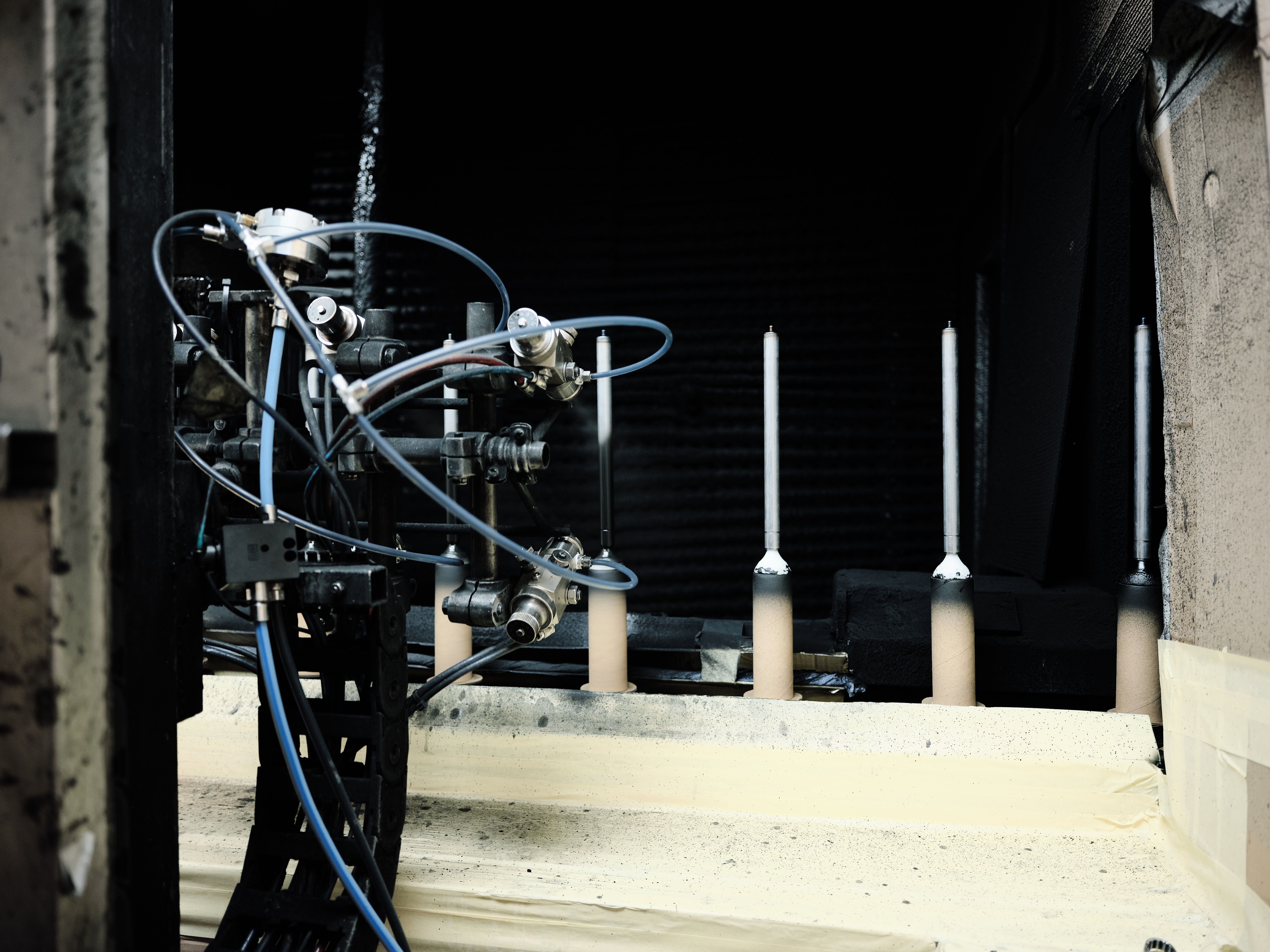 Ein komplexer Roboterarm mit Schläuchen und Kabeln befindet sich in einer schwach beleuchteten industriellen Umgebung. Im Hintergrund sind vier zylindrische Stäbe ausgerichtet.