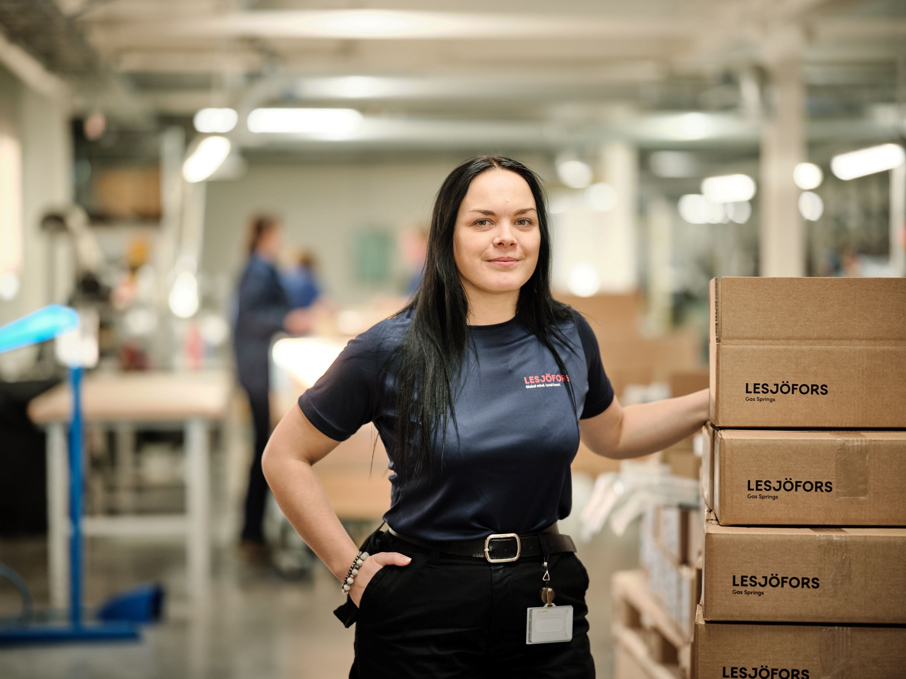 A woman in a blue shirt with Lesjöfors logo stands confidently next to stacked boxes in a well-lit warehouse.