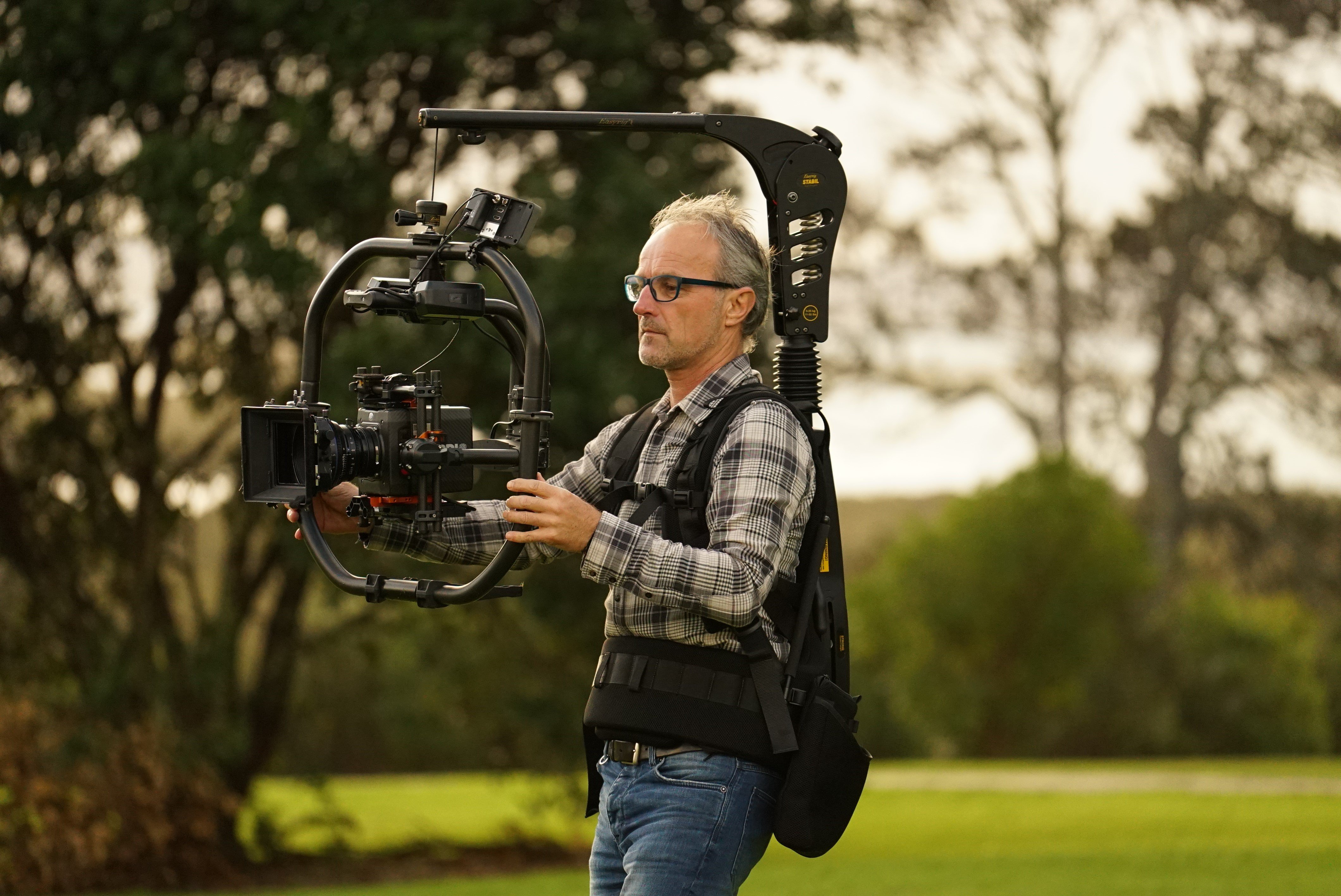 Cameraman wearing a body-mounted stabilizer rig while filming with a professional camera in a grassy outdoor environment.