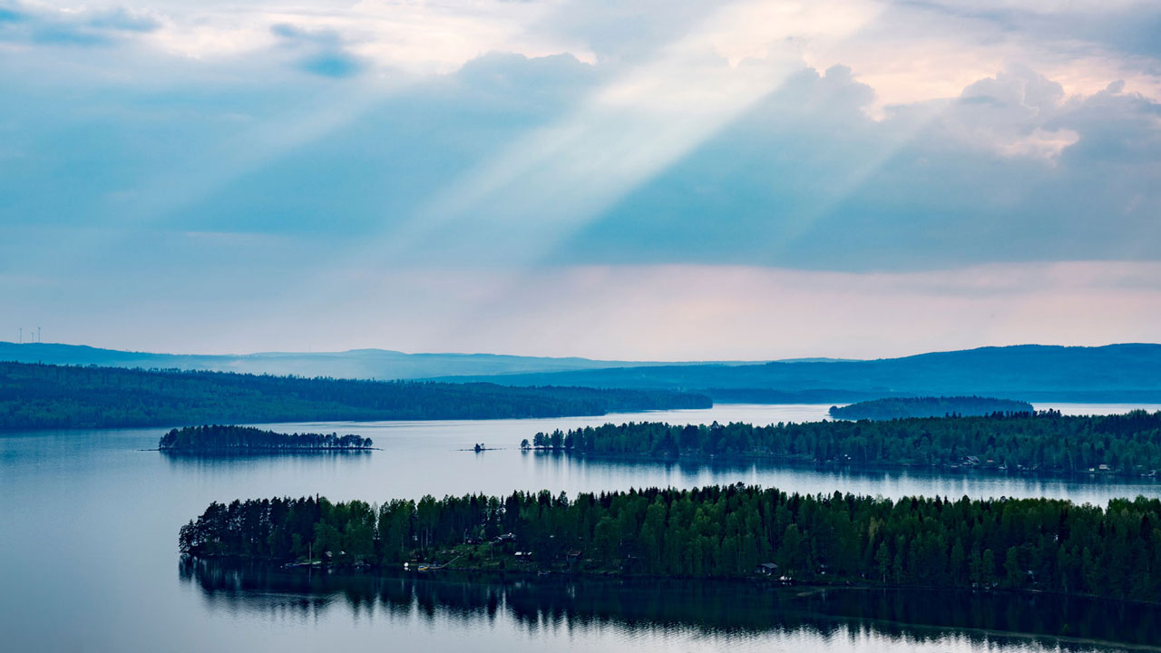 Sun rays breaking through clouds over a lake with forested islands and distant hills.