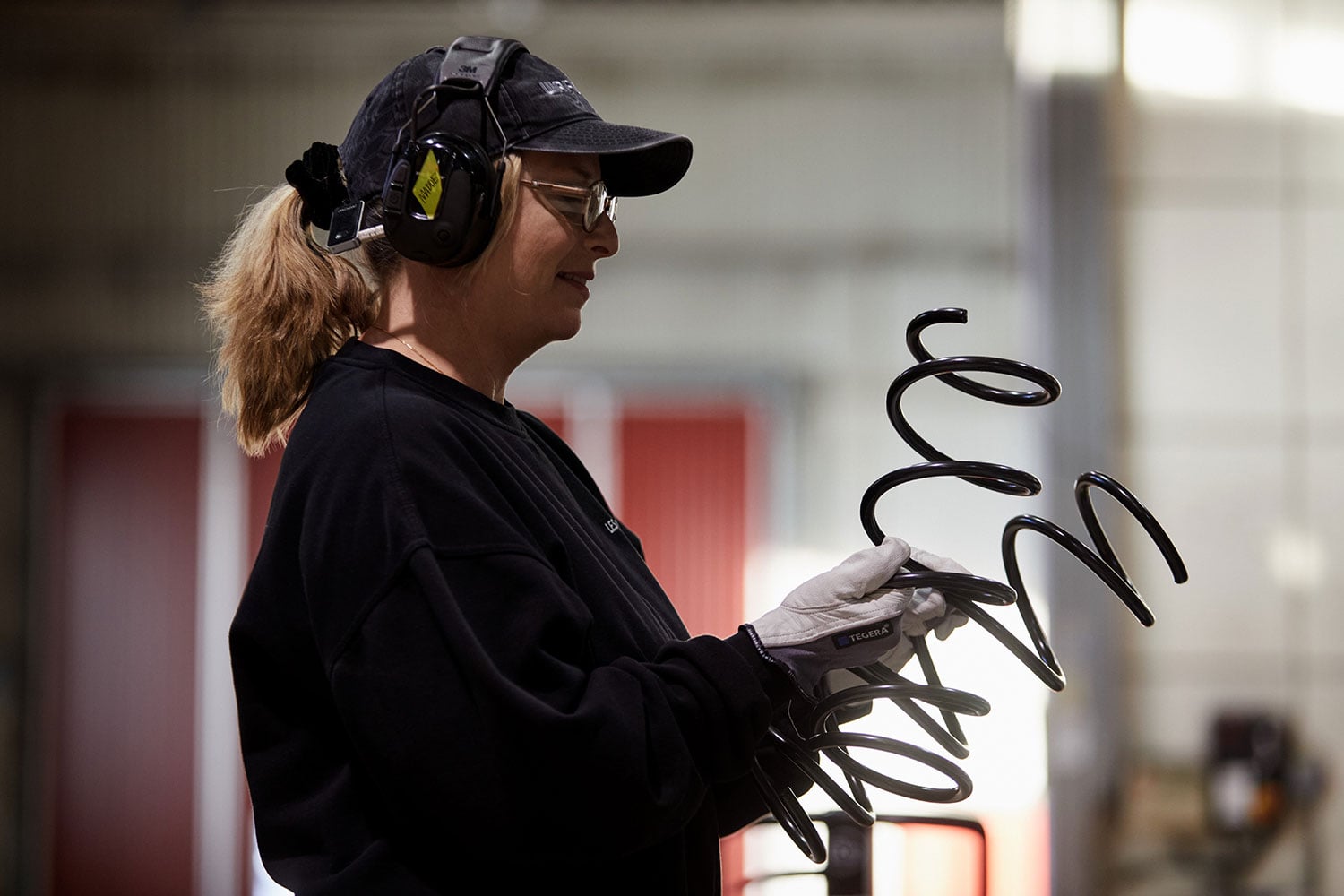 Darbinieks ar cepuri, dzirdes aizsarglīdzekļiem un cimdiem tur un aplūko lielu melnu spirālatsperi, fonā izplūdušas redzamas industriālās iekārtas.